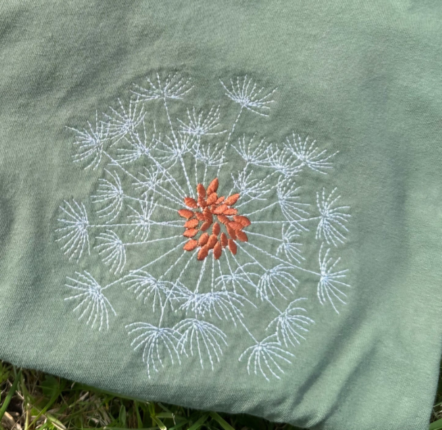 Embroidered dandelion design on a sage  t-shirt with grass in the background