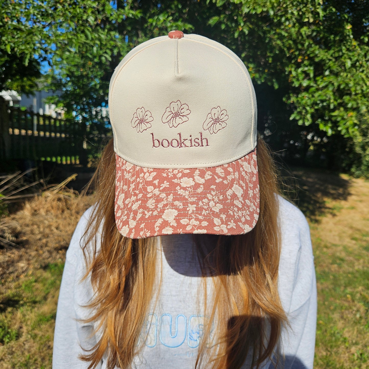 Person wearing a cap with 'bookish' text and floral designs outdoors.