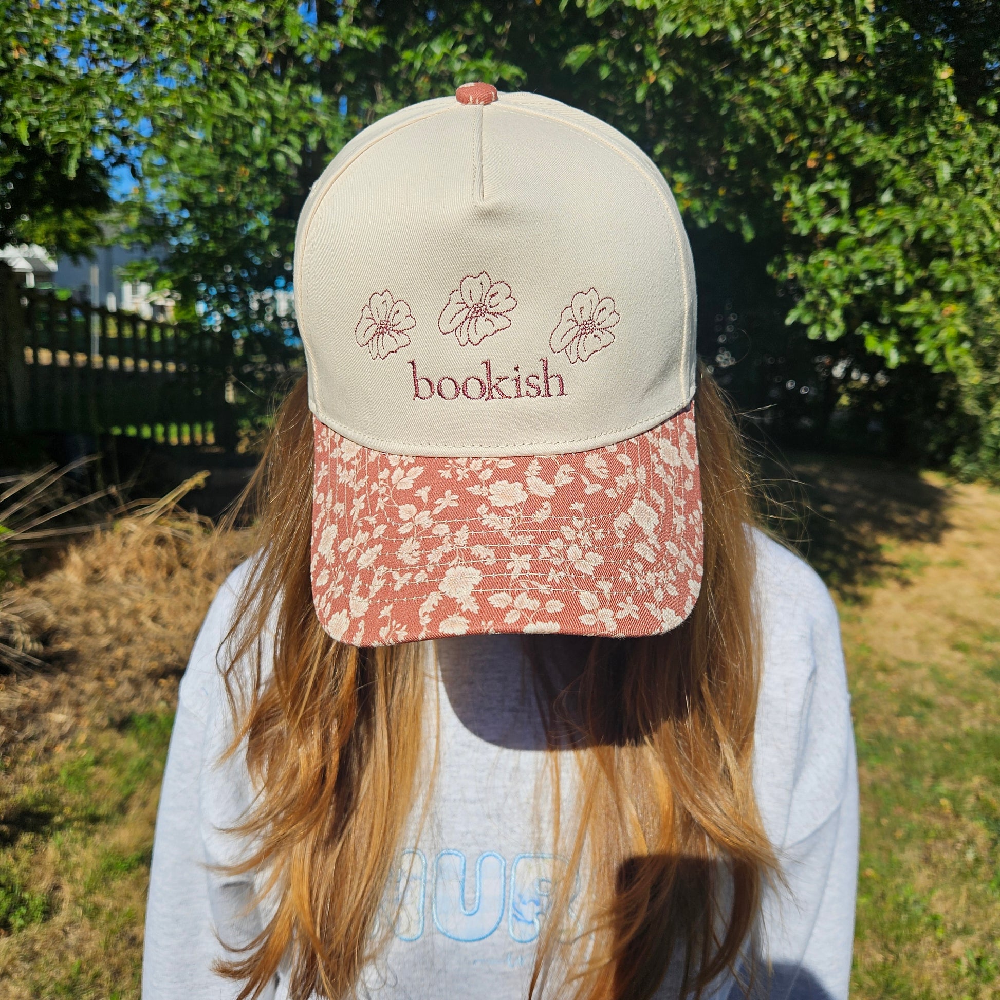 Person wearing a cap with 'bookish' text and floral designs outdoors.