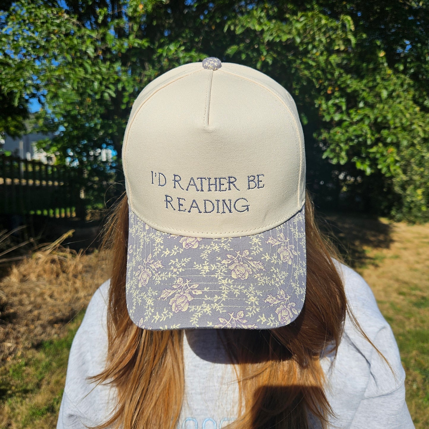 Person wearing a cap with 'I'd Rather Be Reading' text, standing outdoors with trees in the background.