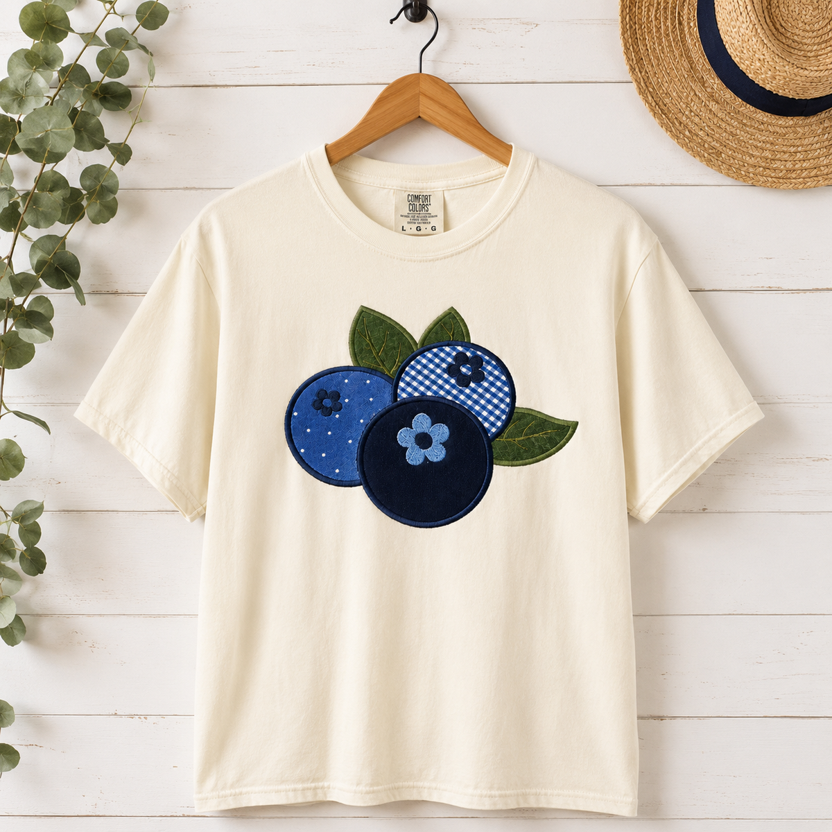 Ivory t-shirt with blue and green floral designs on a wooden surface with a hat and plant.