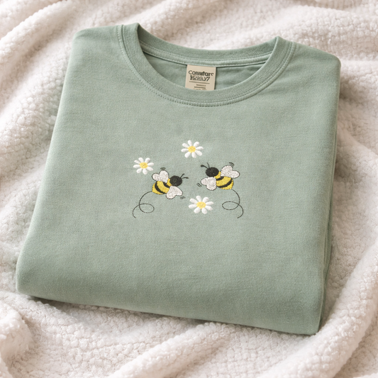 Folded sage t-shirt with bee and daisy design on a soft white background