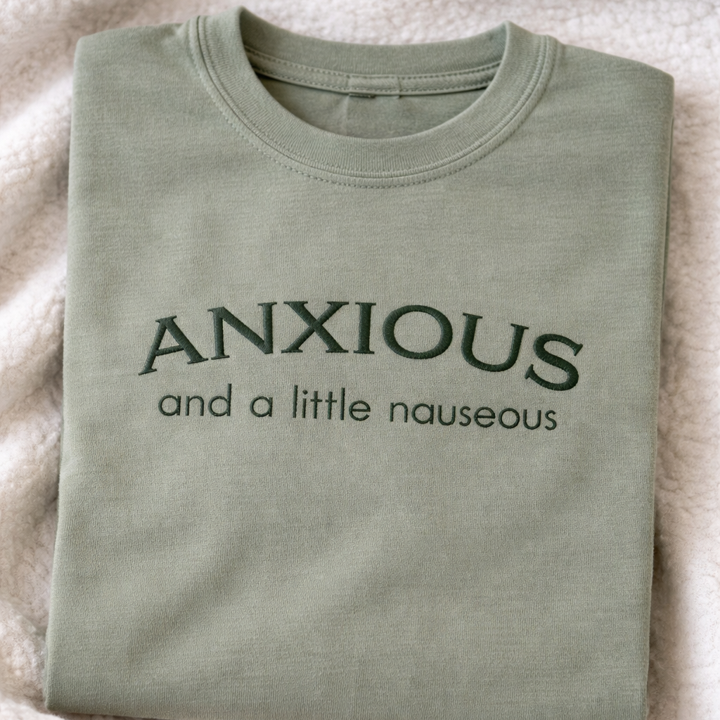 Folded sage t-shirt with embroidered text 'ANXIOUS and a little nauseous' on a white blanket
