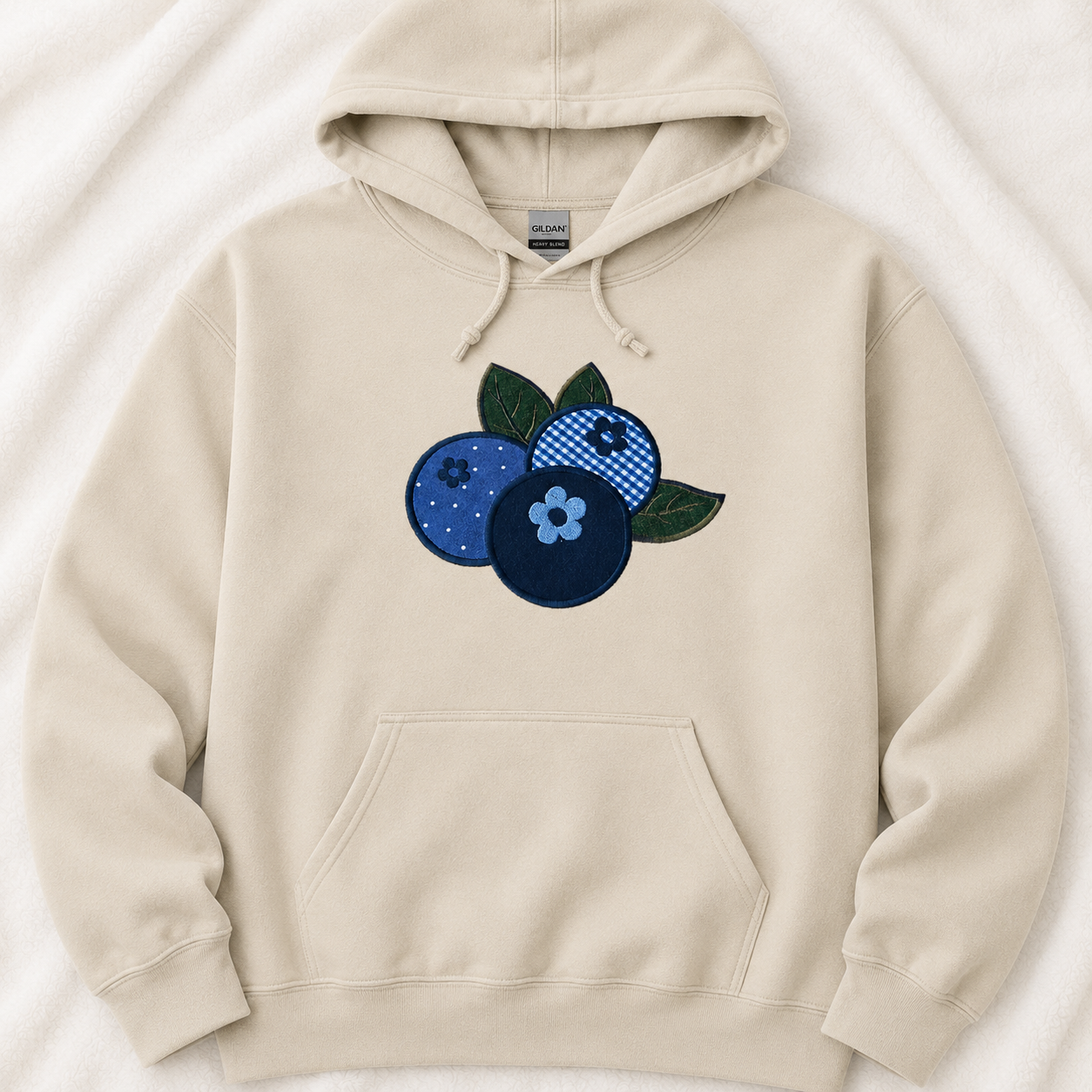 A sand pullover hoodie laid flat on a white textured fabric background, featuring a blueberry applique on the chest with three blueberries in shades of navy, blue, and dark green.